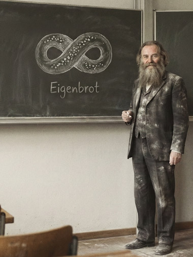 Altes Foto von einem bärtigen Wissenschaftler naben einer Kreidetafel. Er trägt einen Anzu, der mit Kreide- oder Mehl beschmutzt ist. Das gezeichnete Brot sieht aus wie ein Unendlichkeit-Symbol mit Salz